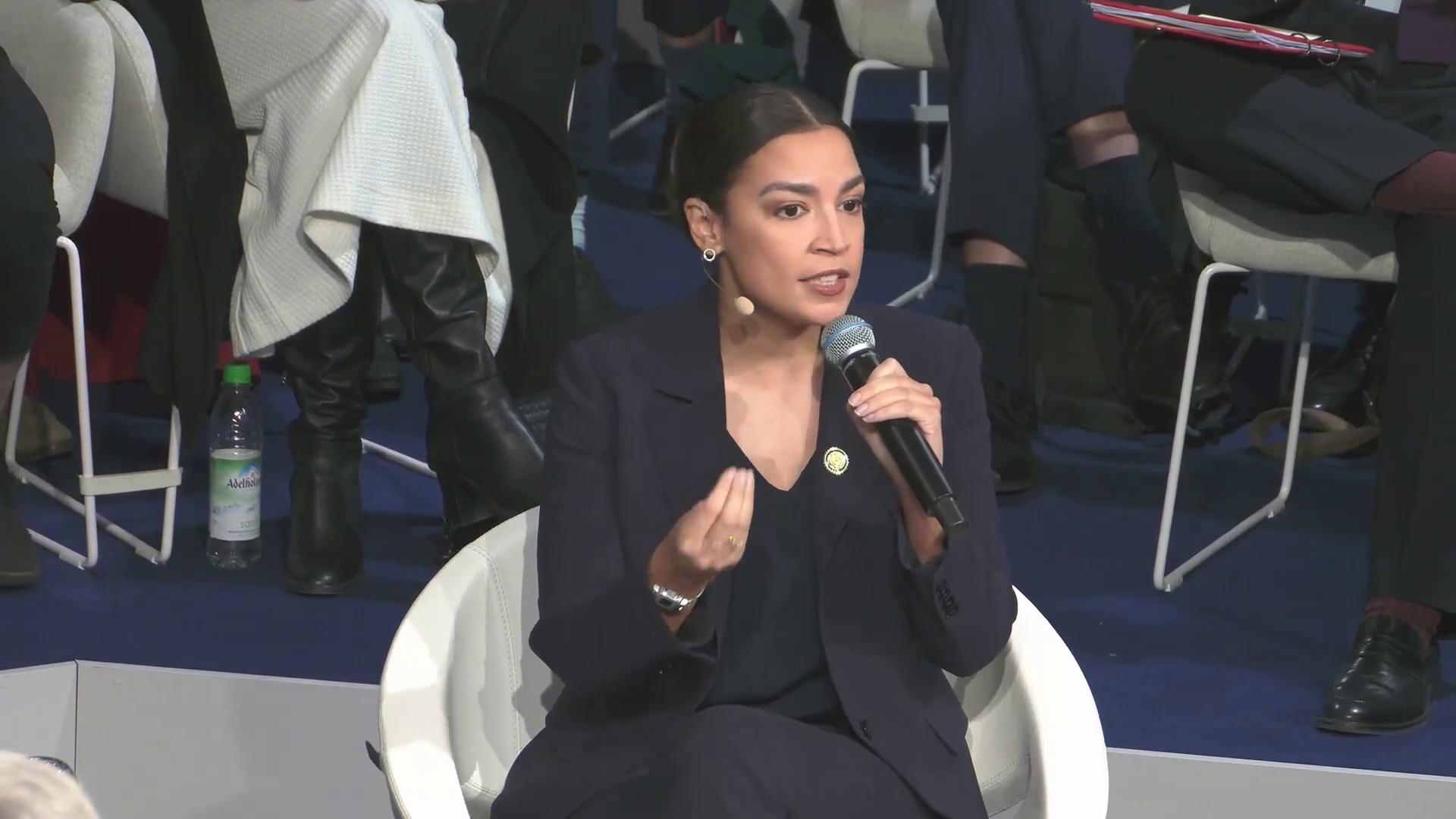 a woman in a suit sitting on a chair alexandria ocasio cortez aoc alexandria ocasiocortez alexandria ocasio cortez sharp and coherent extremely clear and coherent imet2020 beautiful coherent professional film still from dune 2020 alexandrias genesis power pose deepfake