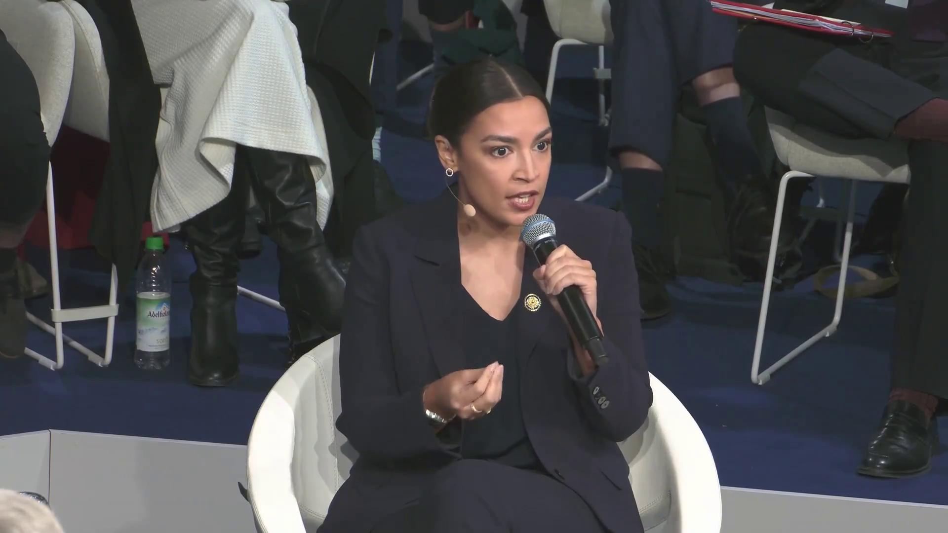 a woman in a suit sitting on a chair alexandria ocasio cortez aoc alexandria ocasiocortez alexandria ocasio cortez sharp and coherent film still from dune 2020 imet2020 extremely clear and coherent beautiful coherent professional totalitarian socialist angel power pose giving a speech deepfake more coherent