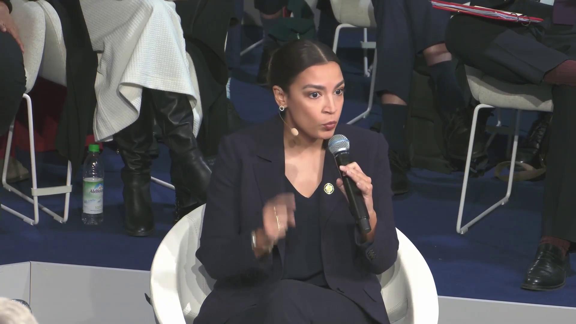 a woman in a black suit sitting on a chair alexandria ocasio cortez aoc alexandria ocasio cortez alexandria ocasiocortez sharp and coherent film still from dune 2020 extremely clear and coherent imet2020 beautiful coherent professional totalitarian socialist angel alexandrias genesis