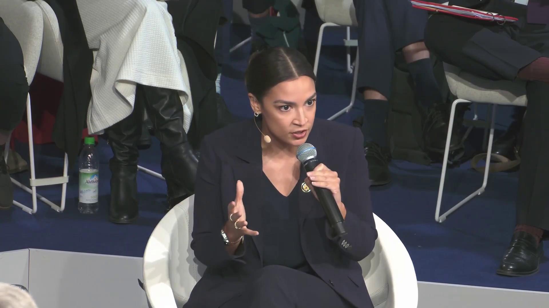 a woman in a black suit sitting on a chair alexandria ocasio cortez alexandria ocasiocortez aoc alexandria ocasio cortez sharp and coherent extremely clear and coherent imet2020 beautiful coherent professional film still from dune 2020 30yearold woman from cuba totalitarian socialist angel