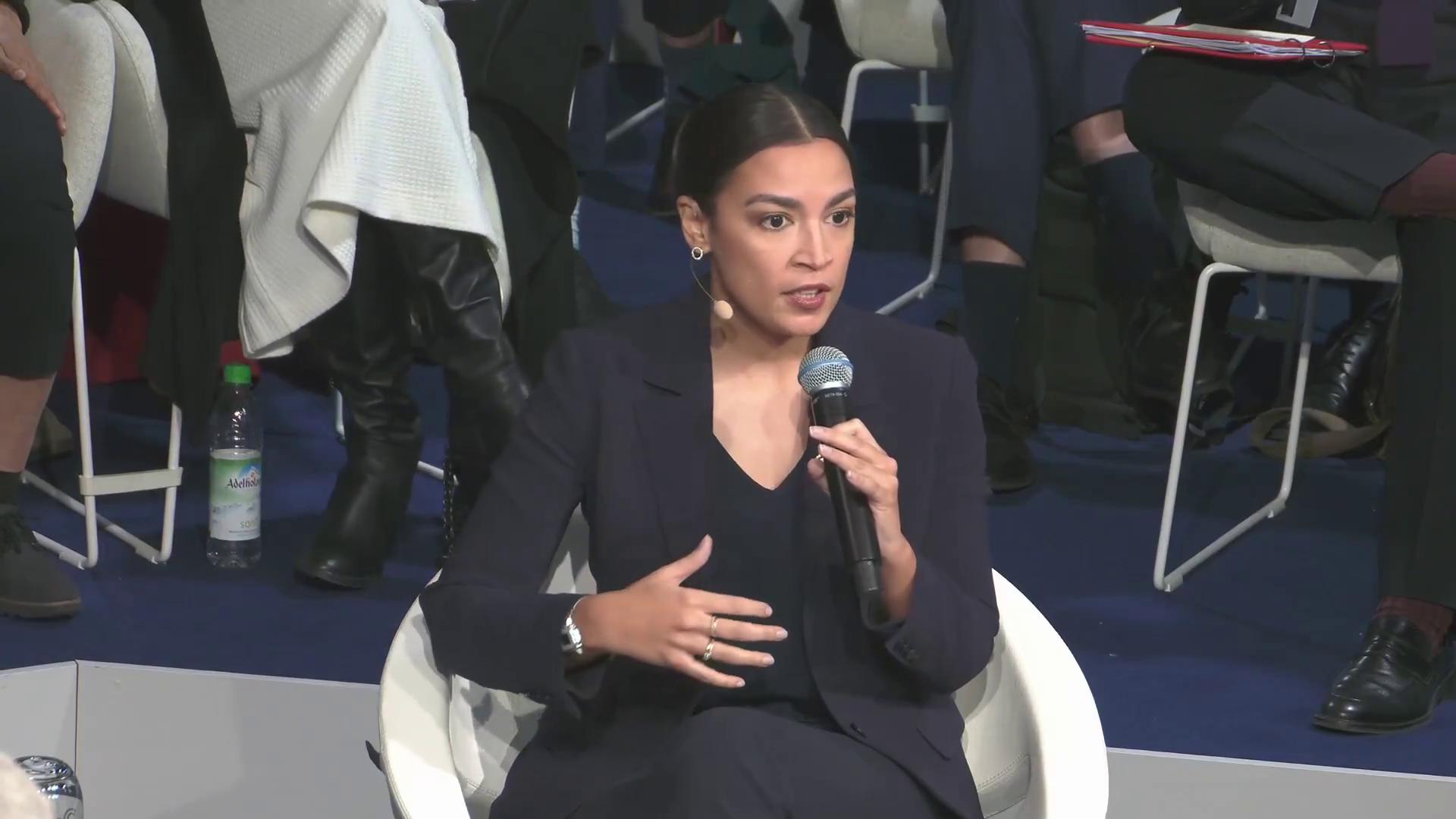 a woman in a suit sitting on a chair alexandria ocasio cortez alexandria ocasiocortez alexandria ocasio cortez aoc sharp and coherent imet2020 extremely clear and coherent film still from dune 2020 beautiful coherent professional alexandrias genesis as a panel of a marvel comic totalitarian socialist angel