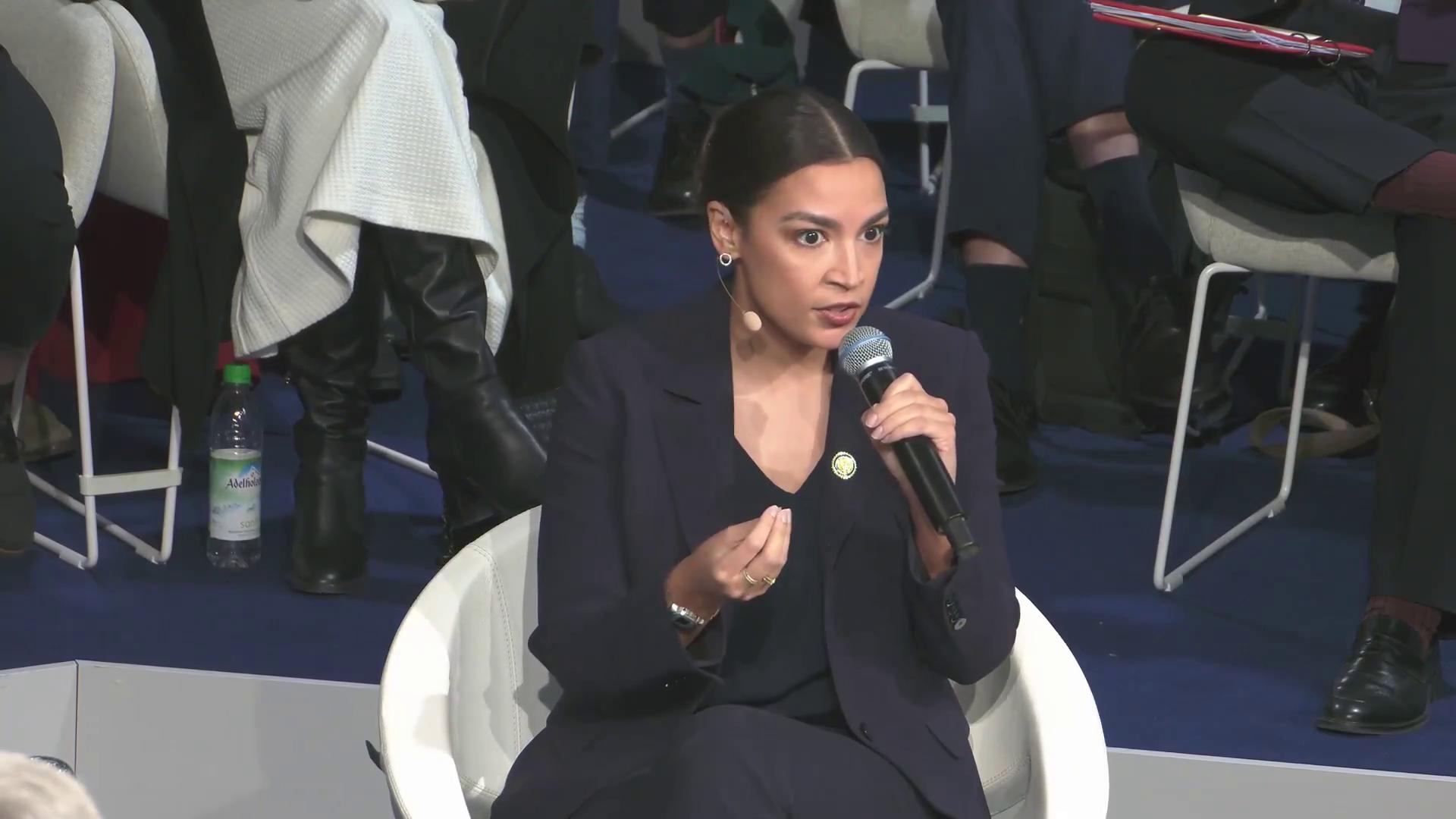 a woman in a suit sitting on a chair alexandria ocasio cortez aoc alexandria ocasiocortez alexandria ocasio cortez sharp and coherent extremely clear and coherent imet2020 beautiful coherent professional film still from dune 2020 alexandrias genesis power pose deepfake