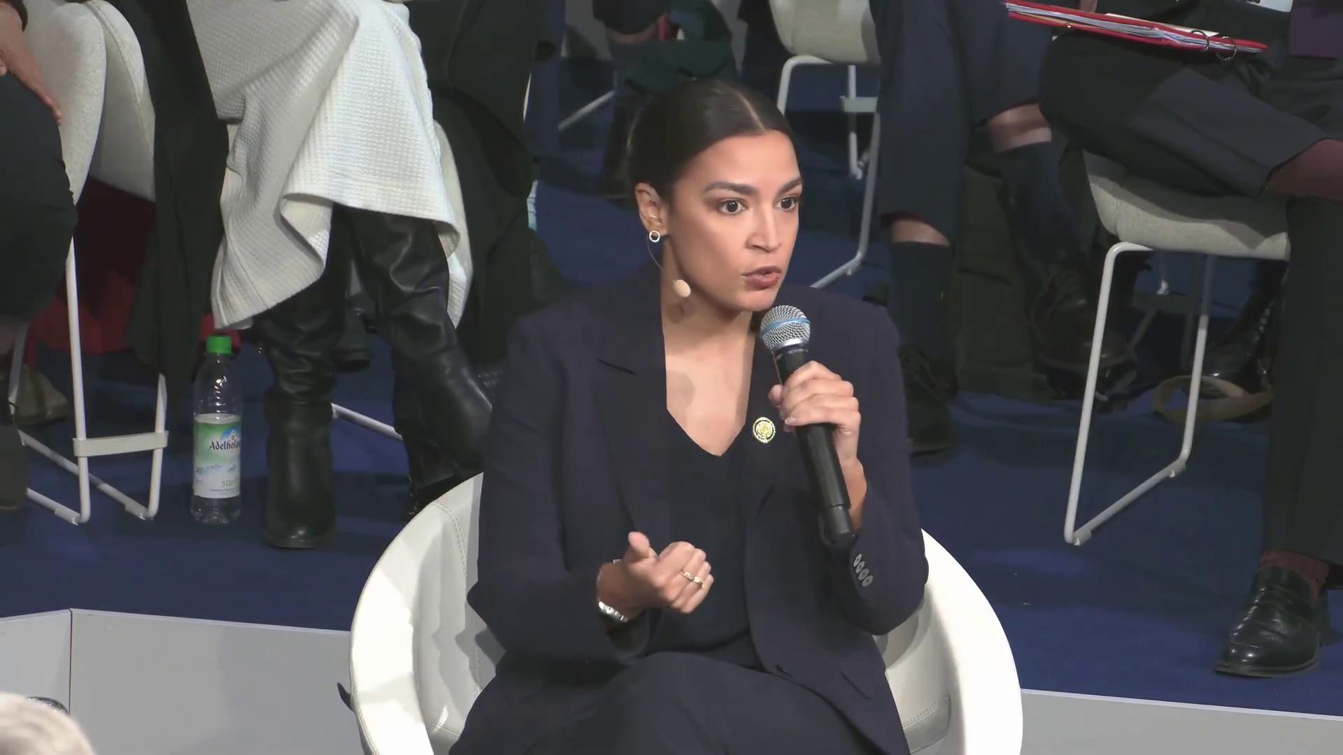 a woman in a black suit sitting on a chair alexandria ocasio cortez aoc alexandria ocasio cortez alexandria ocasiocortez sharp and coherent film still from dune 2020 extremely clear and coherent imet2020 beautiful coherent professional totalitarian socialist angel alexandrias genesis